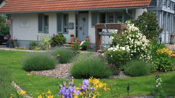 Une maison avec un jardin à l’avant, un toit recouvert de tuiles rouges et une enseigne sur la porte.