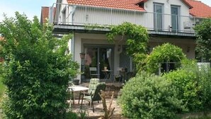 Une maison à deux étages, avec un toit recouvert de tuiles rouges, un balcon et un patio aménagé avec du mobilier d’extérieur.
