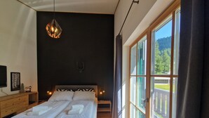 A bedroom with a large bed, a wooden dresser, a wall-mounted TV, and a window with a view of greenery.