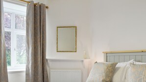 A neatly made bed with pillows and folded towels, a window with curtains, a radiator, and a mirror on the wall. - 3 bedroom accommodation in Arnside, near Grange-over-Sands (Arnside, near Grange-over-Sands)