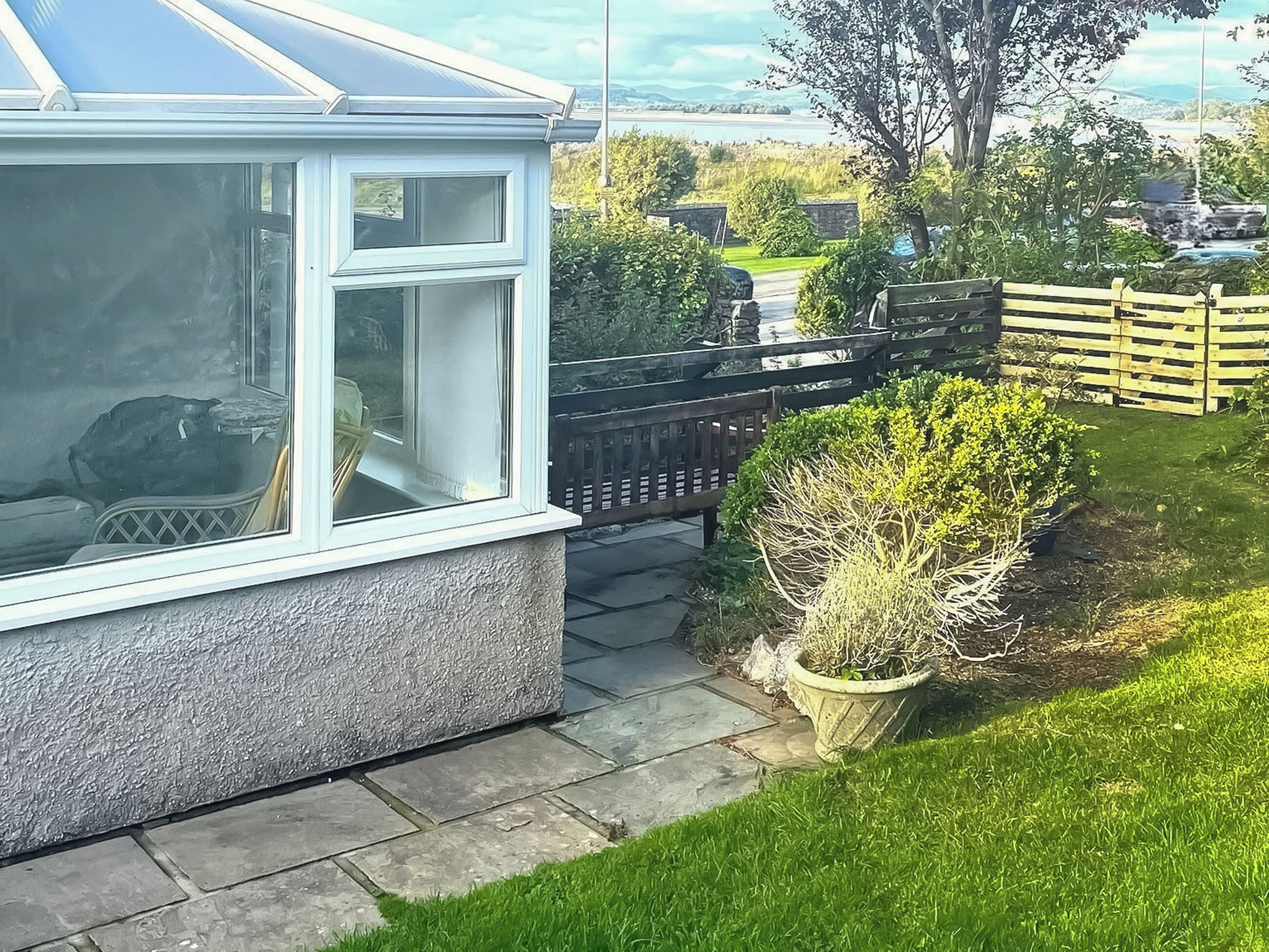 A house with a conservatory, a paved pathway, and a garden with a wooden fence and a bush.