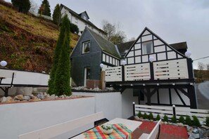 A traditional half-timbered house with a sloped roof, a small garden area with a bench, and a table with a colorful tablecloth.