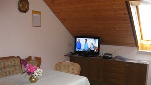 Un coin repas avec une table dressée pour deux personnes, un téléviseur posé sur un meuble en bois et une fenêtre avec des stores.