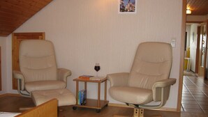 Une pièce chaleureuse avec deux fauteuils beiges, une petite table en bois sur laquelle se trouve un verre de vin rouge et une bibliothèque.