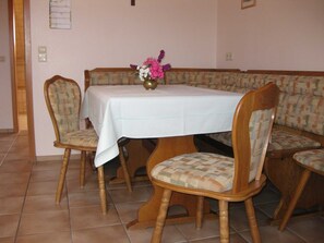 Un coin repas avec une table recouverte d’une nappe blanche, entourée de chaises et de bancs en bois agrémentés de coussins à motifs.