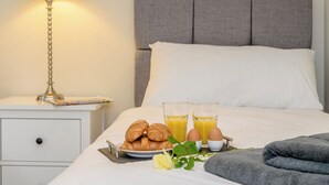 Un lit bien fait, avec une tête de lit grise, une table de chevet avec une lampe, et un plateau de petit-déjeuner comprenant des croissants, des œufs et du jus d’orange.