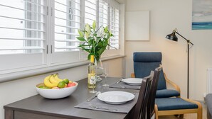 Un coin repas avec une table mise pour deux, un saladier de fruits, un vase avec des fleurs et une chaise.