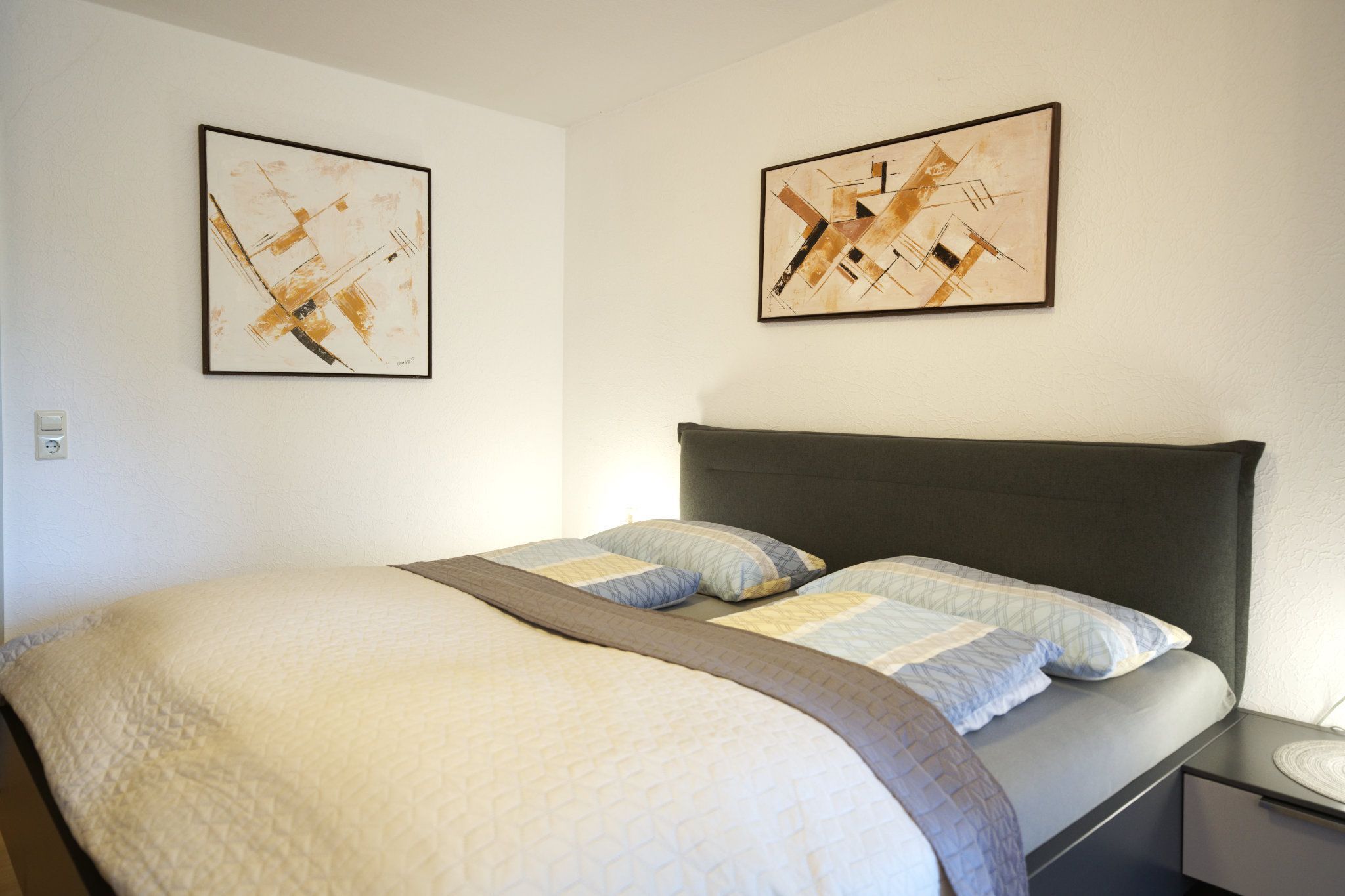 Een hotelkamer met een bed, twee ingelijste abstracte schilderijen, een nachtkastje en een bijzettafeltje.