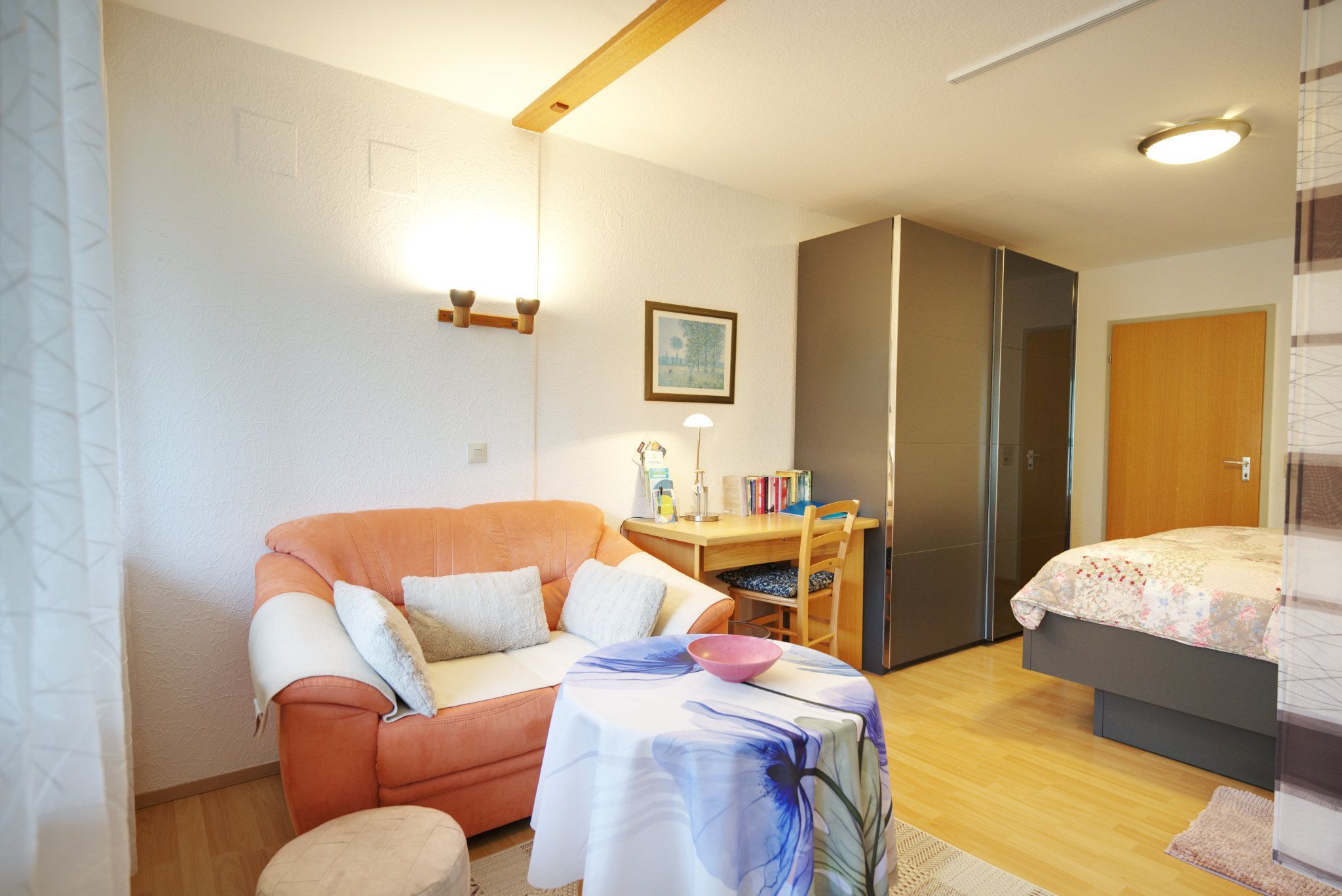 A compact living space with a sofa, a small table with a bowl, a desk with books, a bed, and a door.
