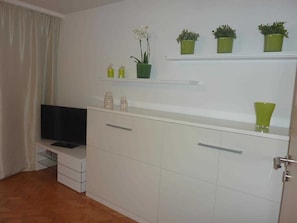 Una habitación moderna y minimalista con muebles blancos, un televisor de pantalla plana y plantas en macetas verdes sobre estantes.