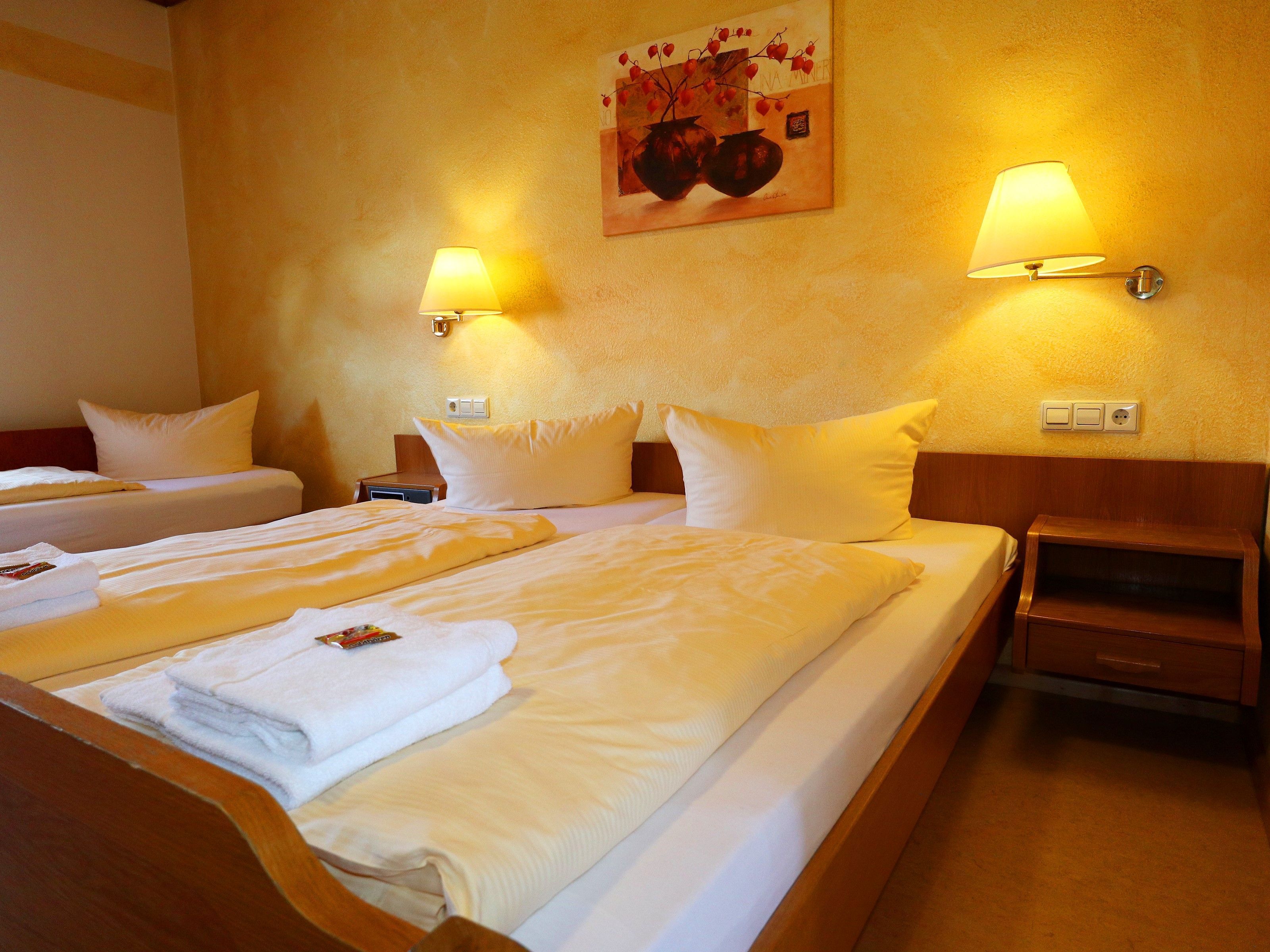 Ein Hotelzimmer mit zwei Betten, jeweils mit weißen Leinen und Kissen, einem Holfnachtkästchen, einer Wandlampe und einem Gemälde über den Betten.