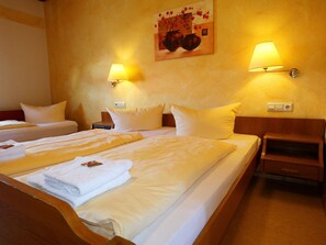 Habitación de hotel con dos camas, cada una con sábanas y almohadas blancas, una mesita de noche de madera, una lámpara de pared y un cuadro sobre las camas.
