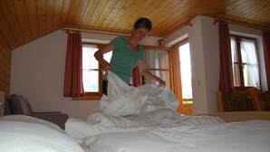 A room with a wooden ceiling, a bed with white bedding, a chair, and a window with curtains.
