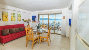 Uma sala de estar com um sofá vermelho, uma mesa de jantar de vidro e cadeiras, e uma varanda com vista para o mar.