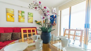 Uma sala de estar moderna com uma mesa de vidro, pratos brancos e uma orquídea em vaso. Há um sofá vermelho e uma cadeira de madeira ao fundo.