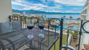 Uma varanda com uma mesa, duas taças de vinho e vista para uma praia e montanhas.