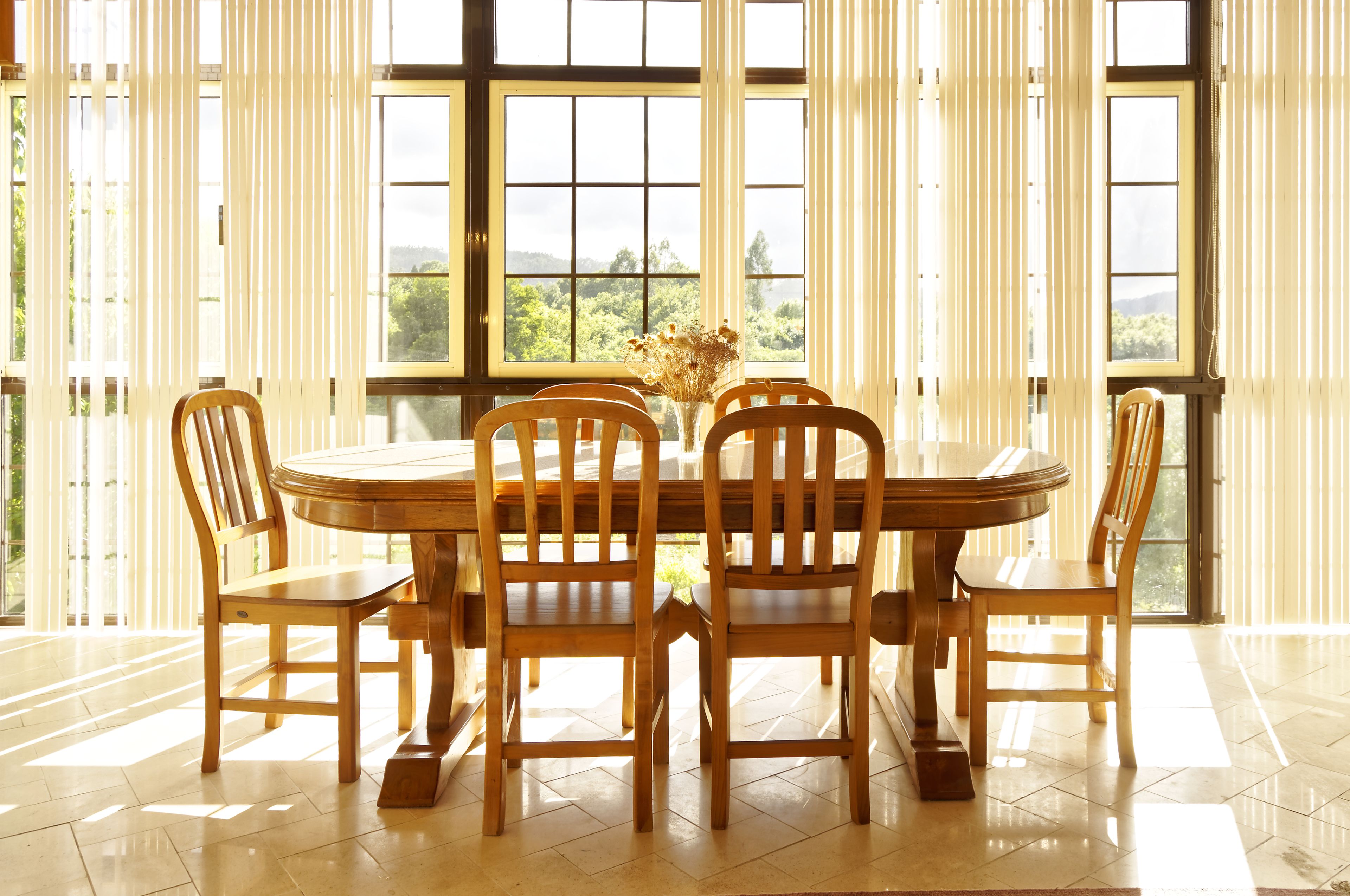 Un comedor con una mesa de madera redonda y seis sillas, ventanas grandes con persianas verticales y vistas al exterior con vegetación.