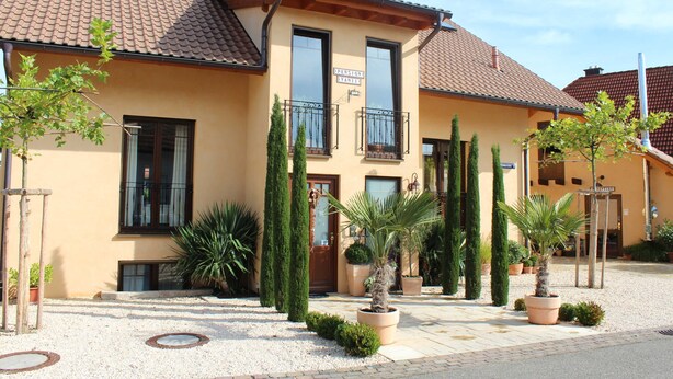 A two-story building with a tiled roof, a balcony with a wrought iron railing, and several potted plants in the front yard. - Two-Bedroom Apartment (4) - Pension Vanii (Rust)
