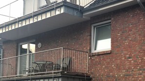 Un bâtiment à deux étages en briques, doté d’un balcon à l’étage supérieur, d’une porte et d’un petit espace extérieur aménagé avec des chaises.