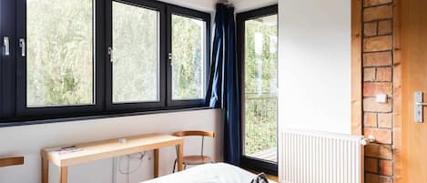 A bedroom with a bed, a desk, and a chair. There are three windows with curtains, a radiator, and a brick wall.