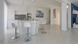 A modern kitchen with white cabinetry, a central island with bar stools, and a painting of a sailboat. - Villa Compass Luxury in a natural bay (Rogoznica)