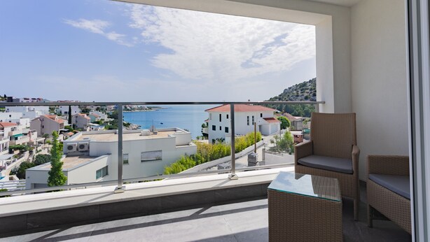 A balcony with wicker furniture, a glass table, and a view of buildings and water. - Villa Compass Luxury in a natural bay (Rogoznica)