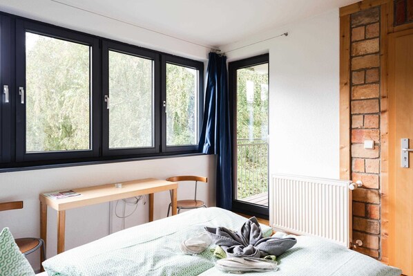 A bedroom with a bed, a desk, and a chair. There are three windows with curtains, a radiator, and a brick wall.