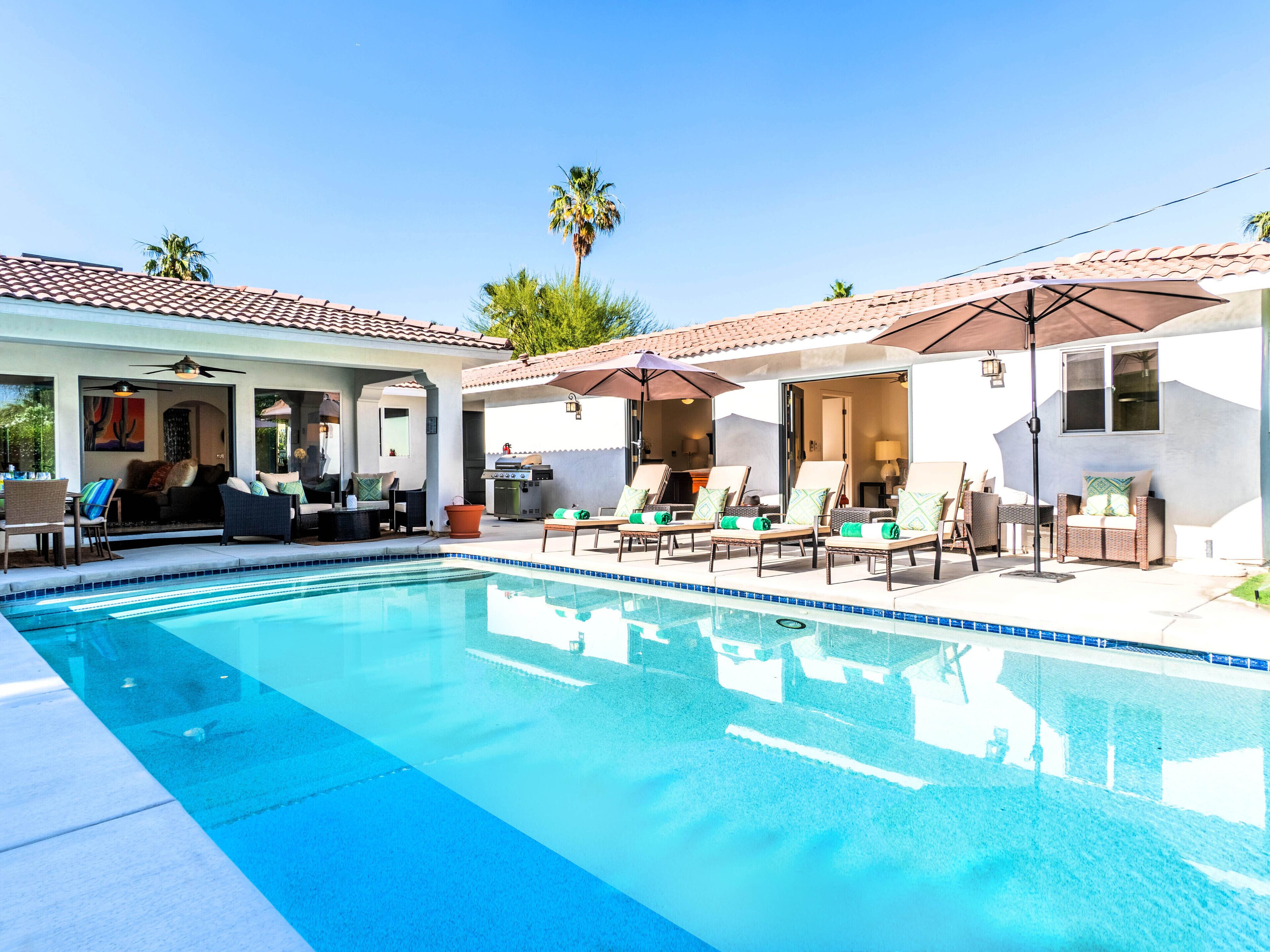 A pool area with lounge chairs and umbrellas, a patio with outdoor furniture, and a building with a tiled roof.