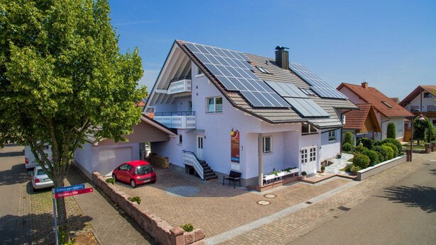 Una casa de dos pisos con paneles solares en el techo, un coche rojo estacionado adelante y un camino de entrada pavimentado.