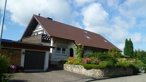 A two-story house with a sloped roof, a stone foundation, and a front garden with flowers and shrubs.