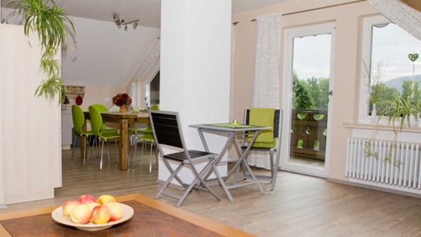 A dining area with a table, chairs, and a bowl of fruit. There is a window with a view of greenery outside.