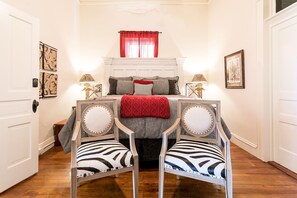 A bedroom with a bed, two chairs, a window with red curtains, and wall art.