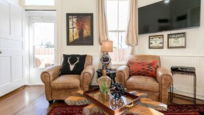 A living room with two leather armchairs, a coffee table, a flat-screen TV, and a framed painting. - In Town Villas | Luxury On Main St. | Hot Tub (Fredericksburg)