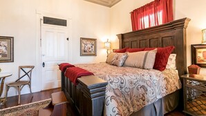A bedroom with a large bed, a wooden headboard, a red curtain, a small table with a lamp, and a chair. - In Town Villas | Luxury On Main St. | Hot Tub (Fredericksburg)