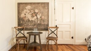 A room with two wooden chairs and a small round table, a large floral painting on the wall, and a white door. - In Town Villas | Luxury On Main St. | Hot Tub (Fredericksburg)