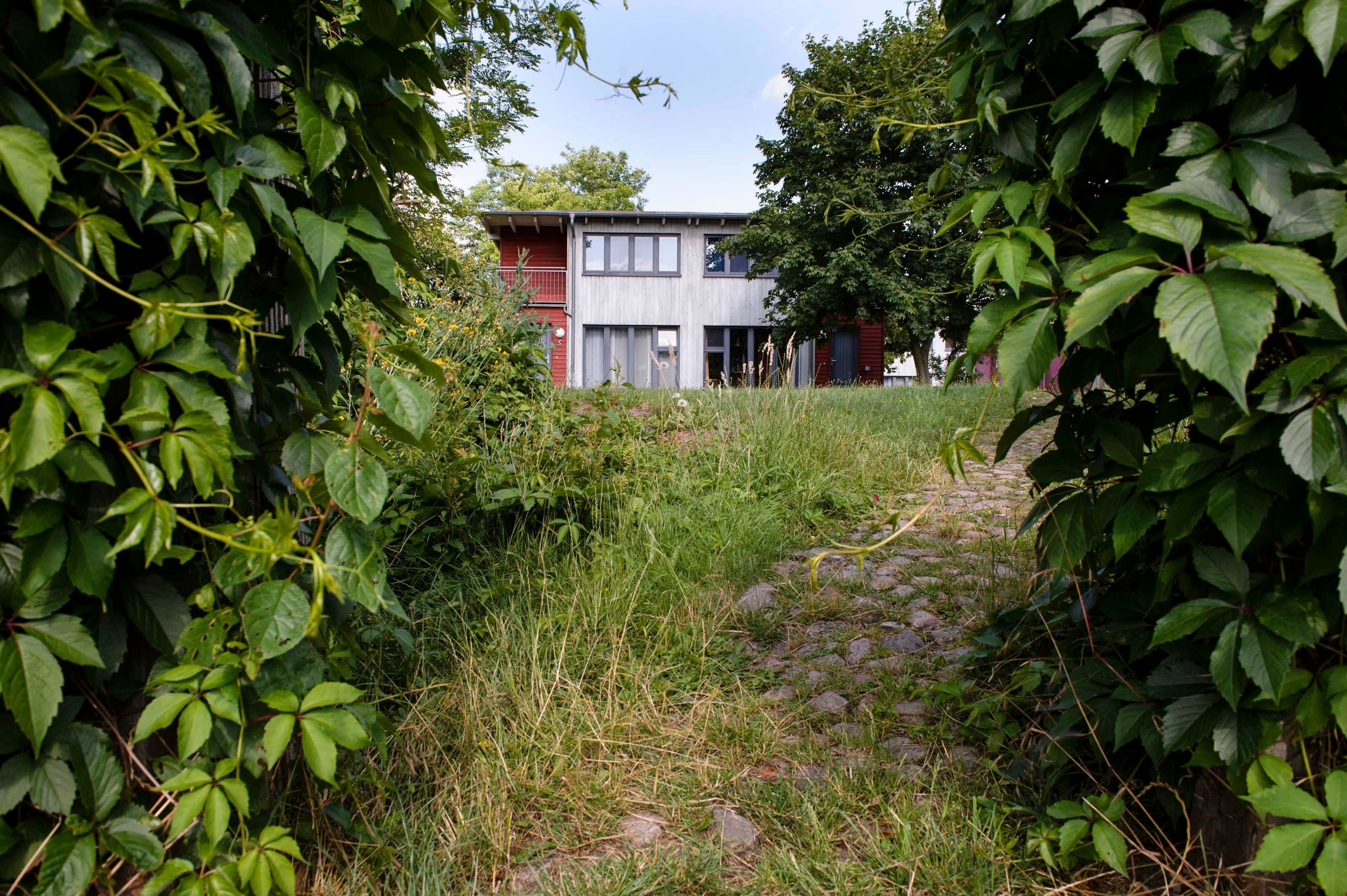 A modern building with a red and grey facade, surrounded by lush greenery and a cobblestone path.
