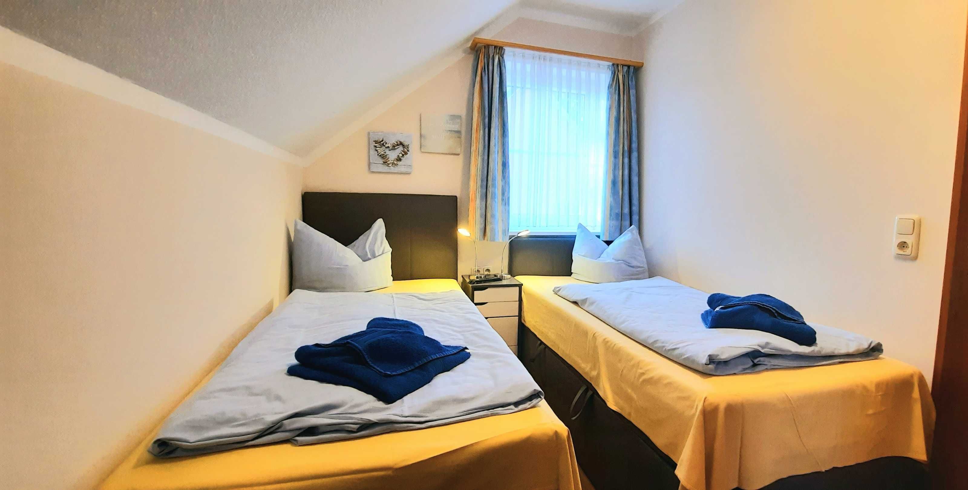 Une petite chambre d’hôtel avec deux lits simples, chacun muni d’un oreiller et d’une serviette pliée. Il y a une table de chevet avec une lampe entre les lits, ainsi qu’une fenêtre avec des rideaux.