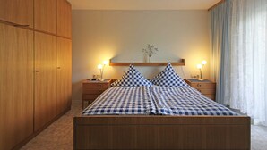 A bedroom with a wooden bed, checkered bedding, two bedside tables with lamps, a nightstand, and a large wardrobe. - Double room blue - Winery Baumer (Vogtsburg im Kaiserstuhl)