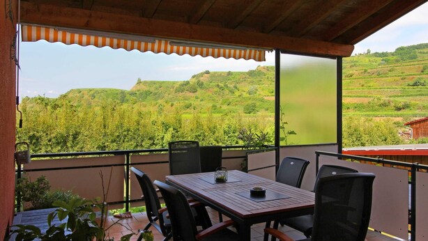 A covered outdoor seating area with a table and chairs, overlooking a lush green landscape with hills and vineyards. - Double room blue - Winery Baumer (Vogtsburg im Kaiserstuhl)