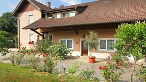 A two-story house with a tiled roof, a front porch, and a garden with various plants and flowers. - Double room blue - Winery Baumer (Vogtsburg im Kaiserstuhl)