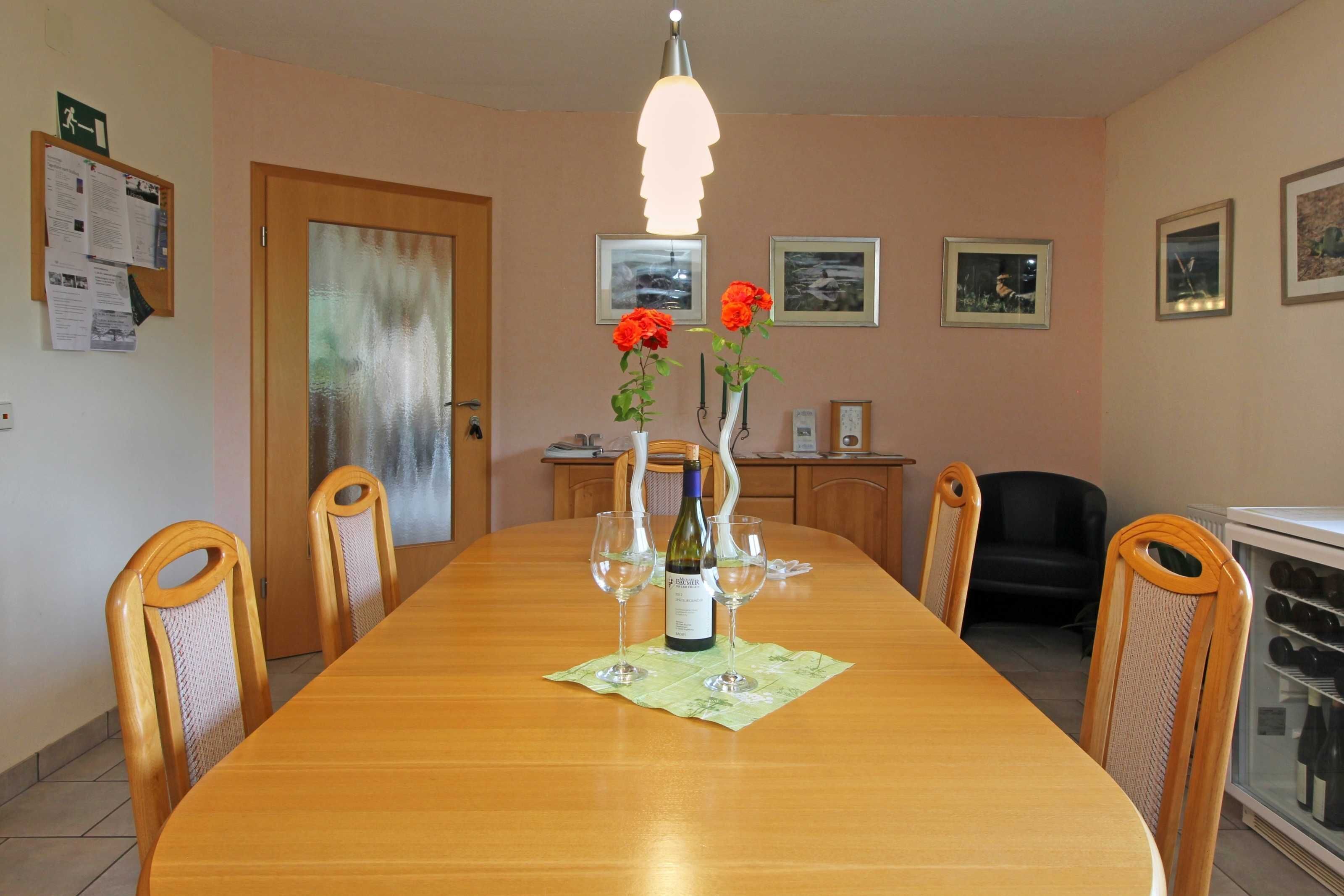 Un comedor con una mesa de madera, sillas, una botella de vino y vasos. Hay cuadros en las paredes y un refrigerador con botellas.