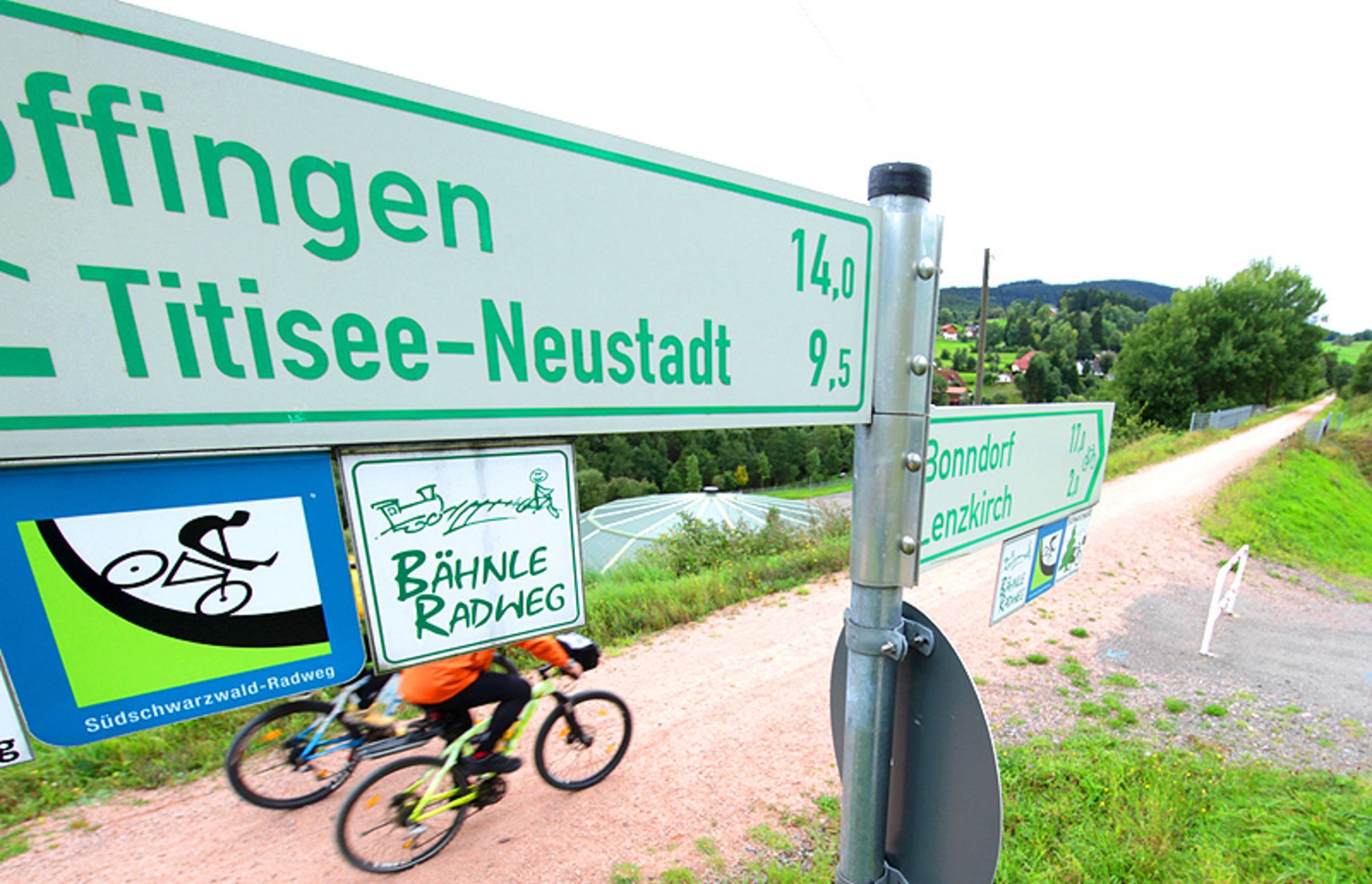 Ein Wegweiser mit Entfernungen zu Städten und ein Radfahrer auf einem Feldweg.