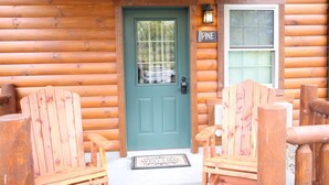 Een houten blokhut met een groene deur, twee Adirondack houten stoelen, een deurmat en een bordje met de tekst 'Pine'.