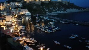 Un pueblo costero al anochecer con una estructura fortificada en la cima de una colina, un puerto deportivo con numerosas embarcaciones y edificios con ventanas iluminadas.