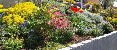 A garden with various colorful flowers and plants, a stone retaining wall, and a building in the background.