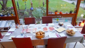 Un espace de restauration en plein air avec une table dressée pour un repas, entourée de chaises, et une vue sur un jardin à travers de grandes fenêtres.