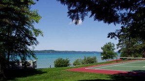 Una cancha de tenis con superficie roja, rodeada de árboles y vista a un lago.