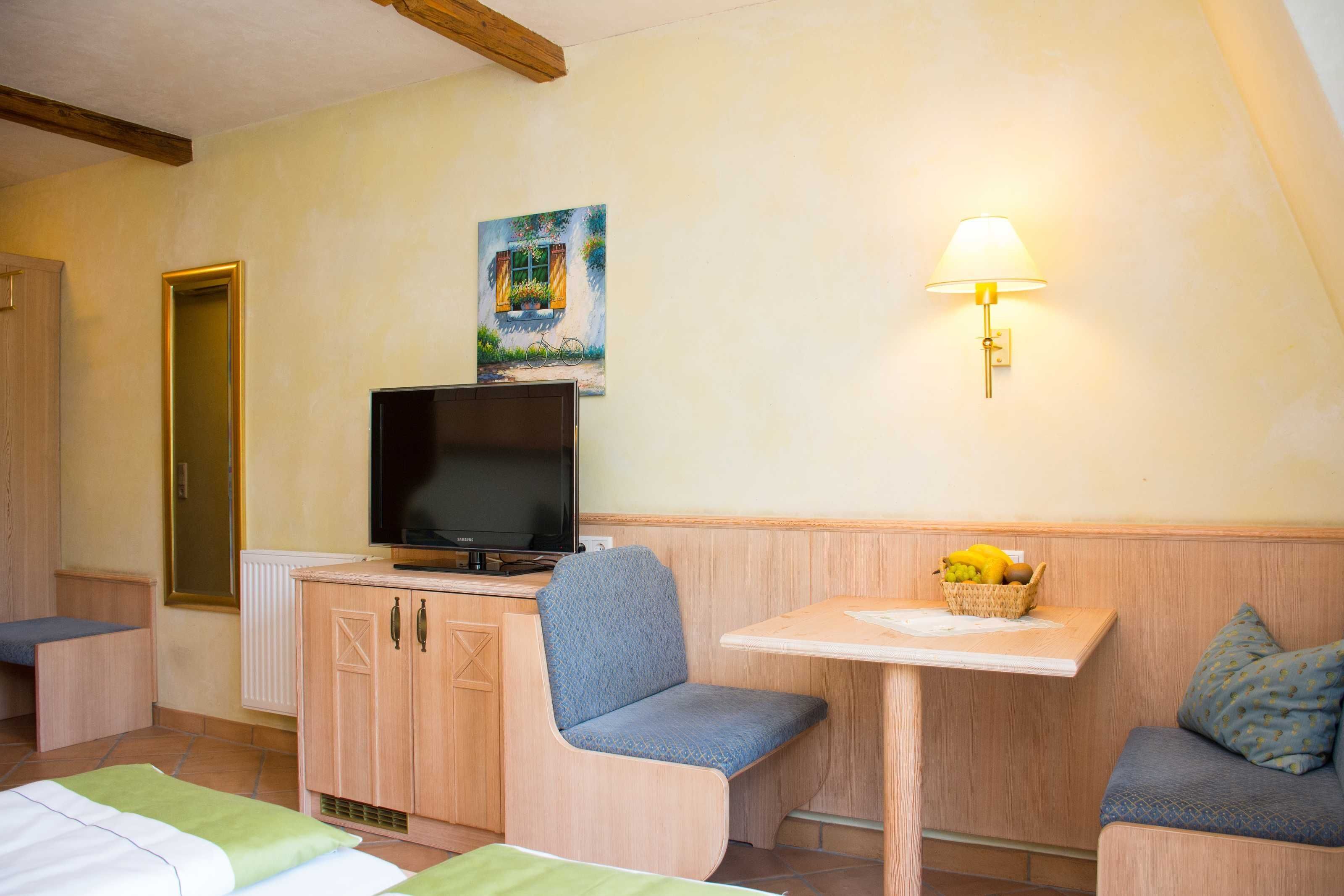 Ein gemütliches Zimmer mit Fernseher auf einer Holzkommode, einem Bild an der Wand, einer Lampe und einem kleinen Tisch mit Obstschale.