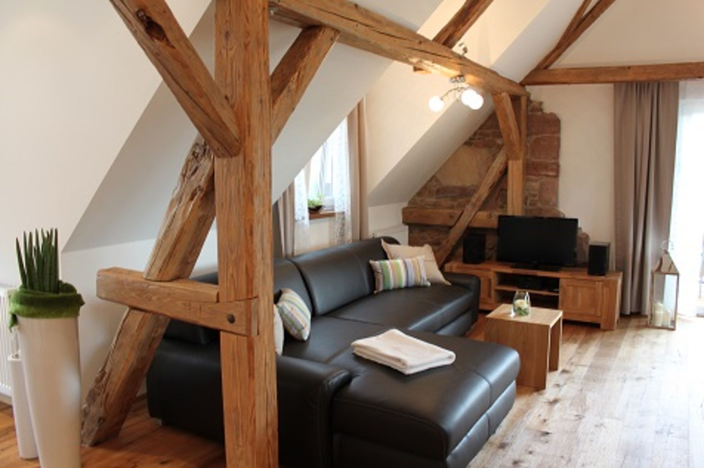 A loft-style living room with exposed wooden beams, a black leather sofa, a television, and a small table.