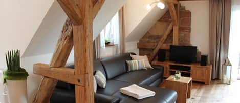 A loft-style living room with exposed wooden beams, a black leather sofa, a television, and a small table.